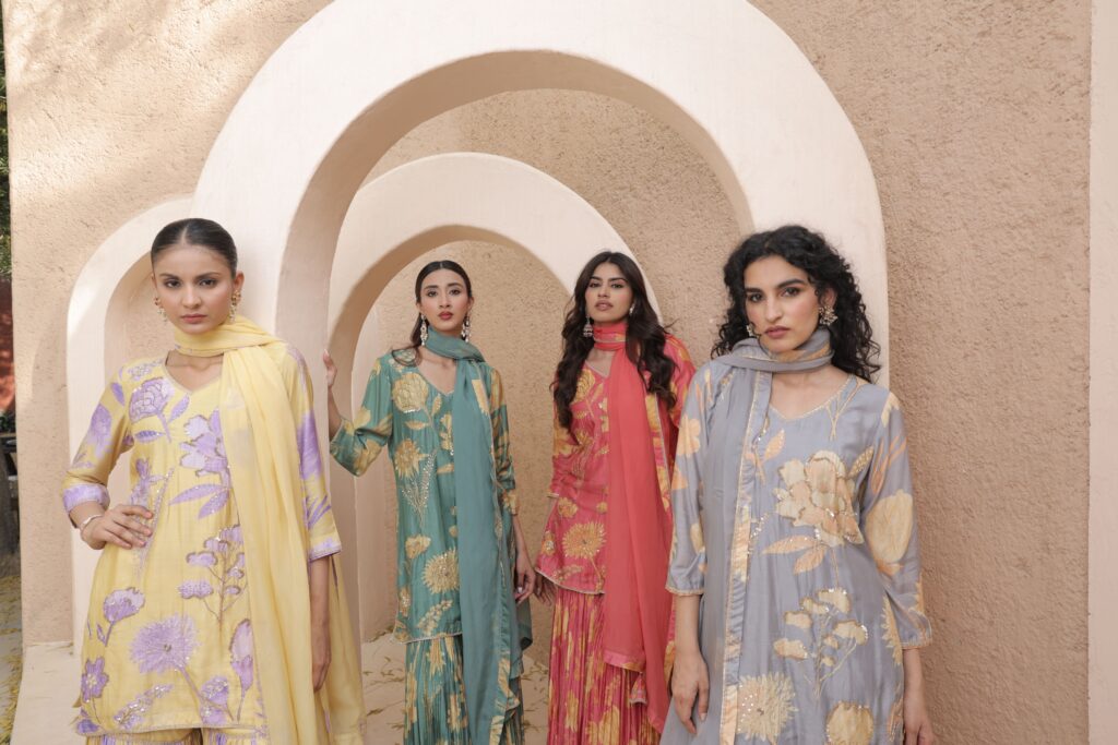 Four women wearing colorful ethnic dresses pose in an elegant, arched setting.